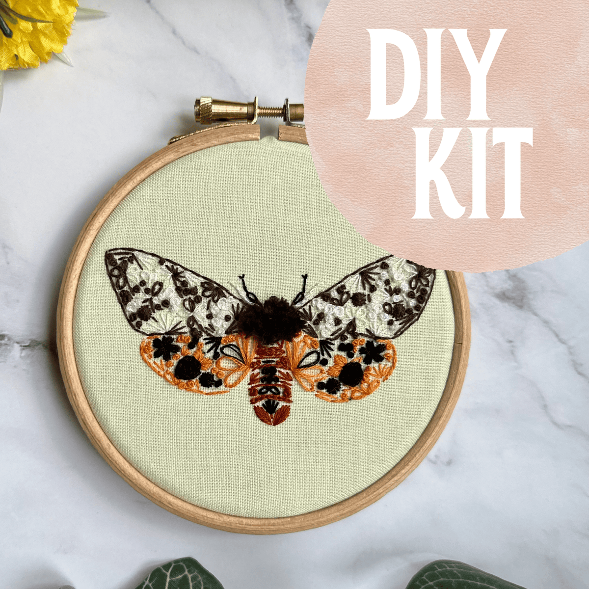 The 4 Beginner Garden Tiger Moth Embroidery Kit by Nest Embirdery features a detailed moth design in a hoop on marble. A pink circle with DIY KIT text is in the corner, and yellow flowers with green leaves are partially visible.