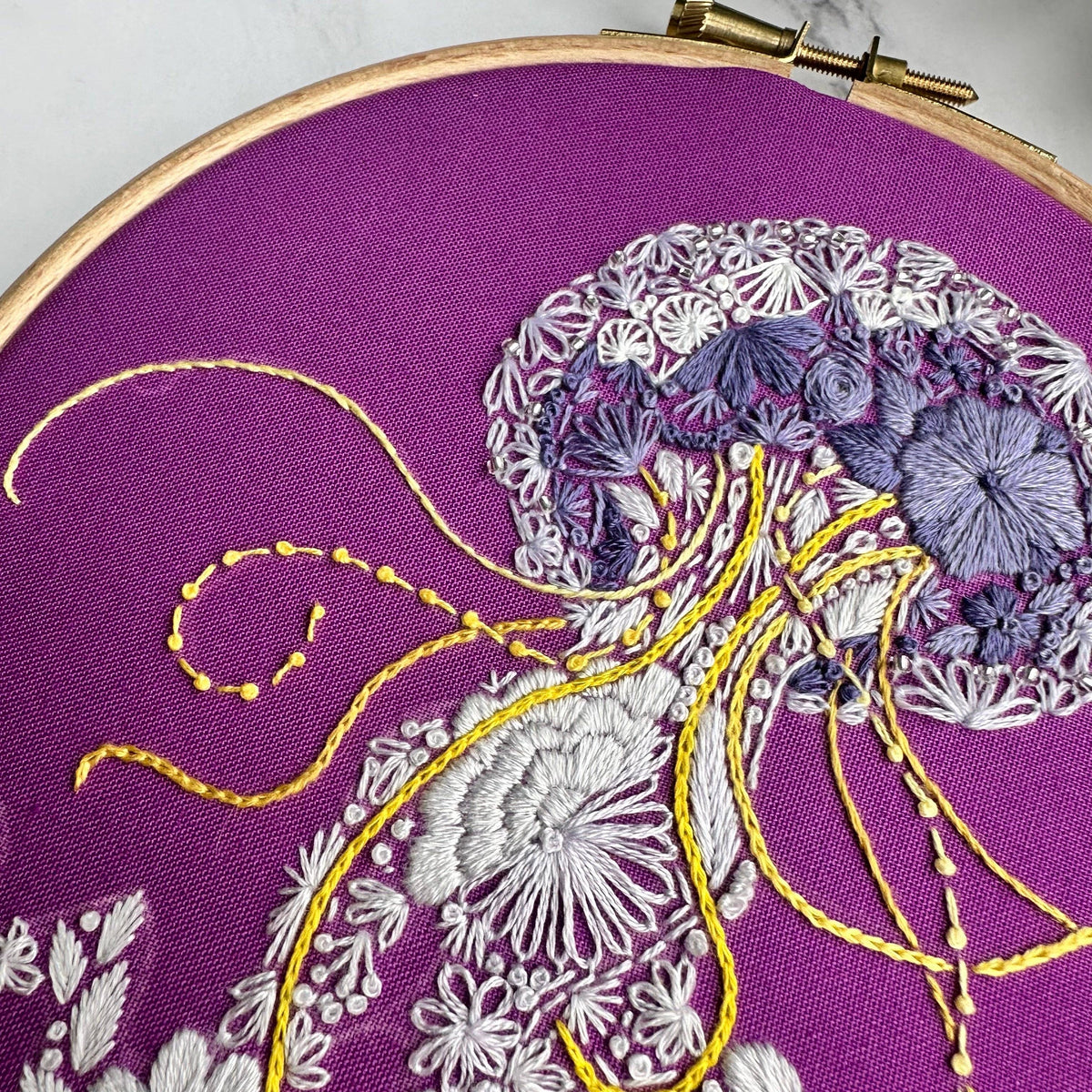 The 6 Beginner Jelly-Stitch Embroidery Kit by Nest Embirdery features a magenta fabric in a hoop with a jellyfish embroidered using white, lavender, and yellow threads. Floral patterns within the jellyfish add an elegant touch for beginners.