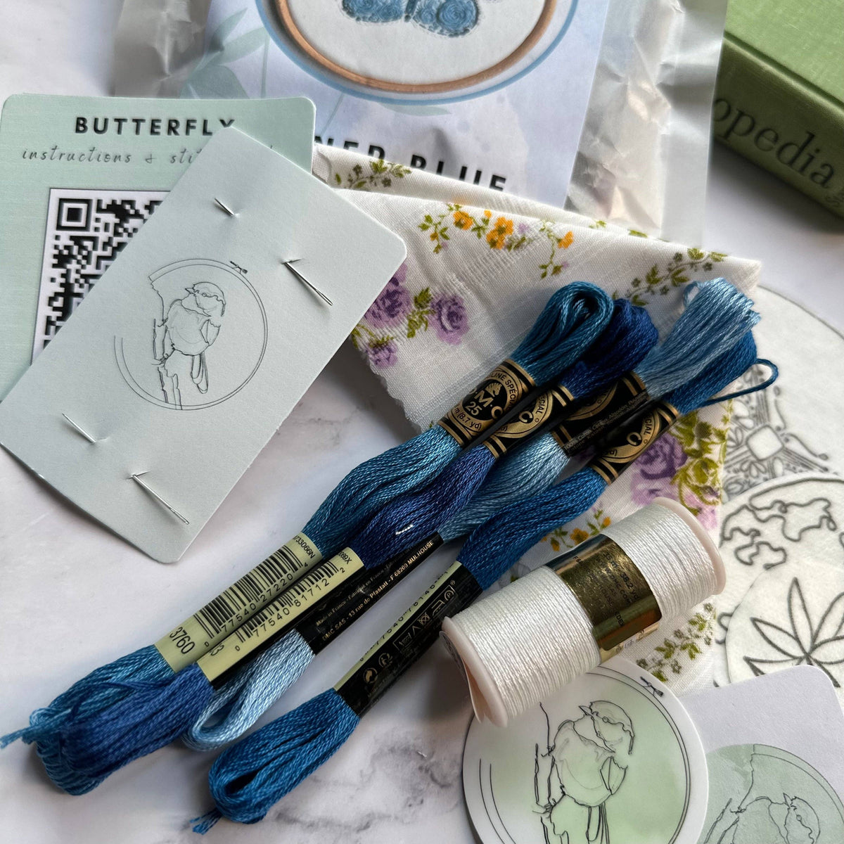 The 4 Beginner Karner Blue Butterfly Embroidery Kit by Nest Embirdery includes blue and white thread, gold spool, floral cloth, needle cards, a woman’s outline sticker, and butterfly packaging—all inspired by the Karner Blue Butterfly.