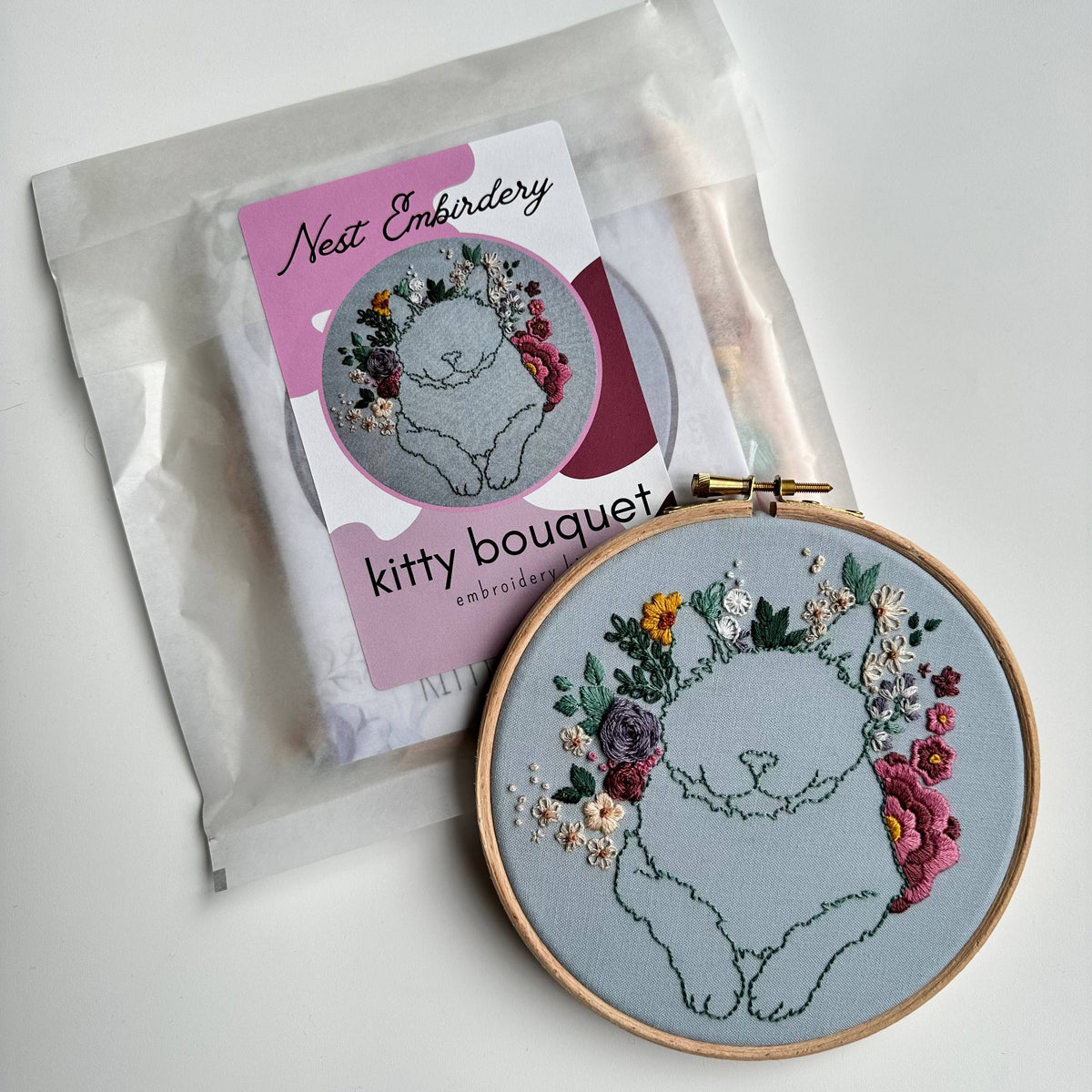 The 6 Beginner Kitty Bouquet Kit from Nest Embirdery features a green-outlined cat surrounded by colorful flowers on light blue fabric—ideal for cat lovers and advanced beginners. Includes a finished hoop with the floral cat design.