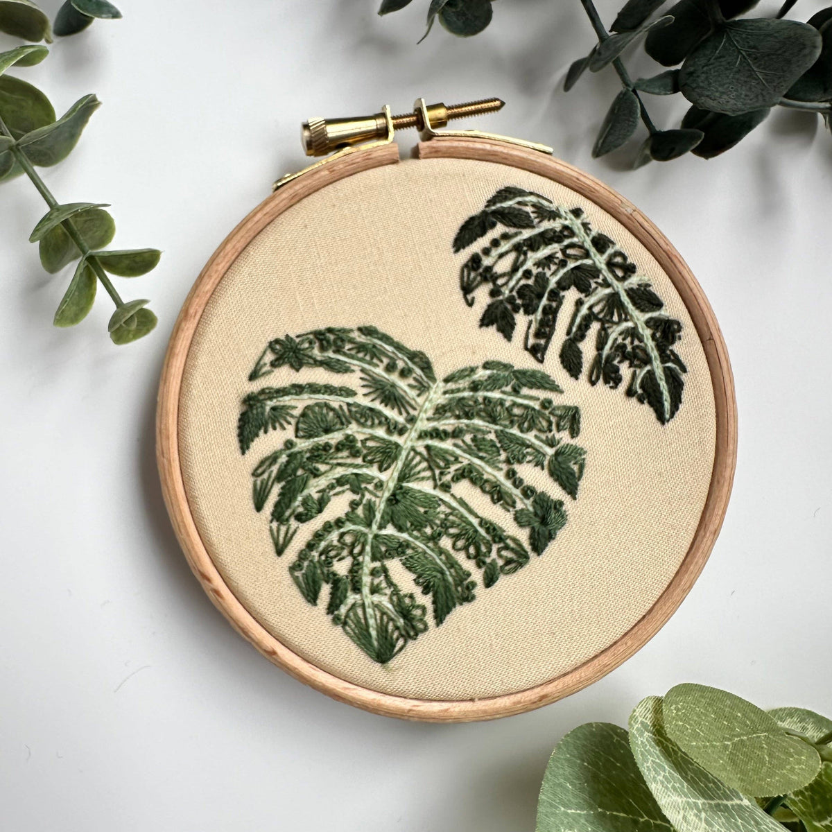 The 4 Beginner Mighty Monstera Embroidery Kit by Nest Embirdery features green monstera leaves stitched on beige fabric in an embroidery hoop, with artificial green leaves arranged on a white background.
