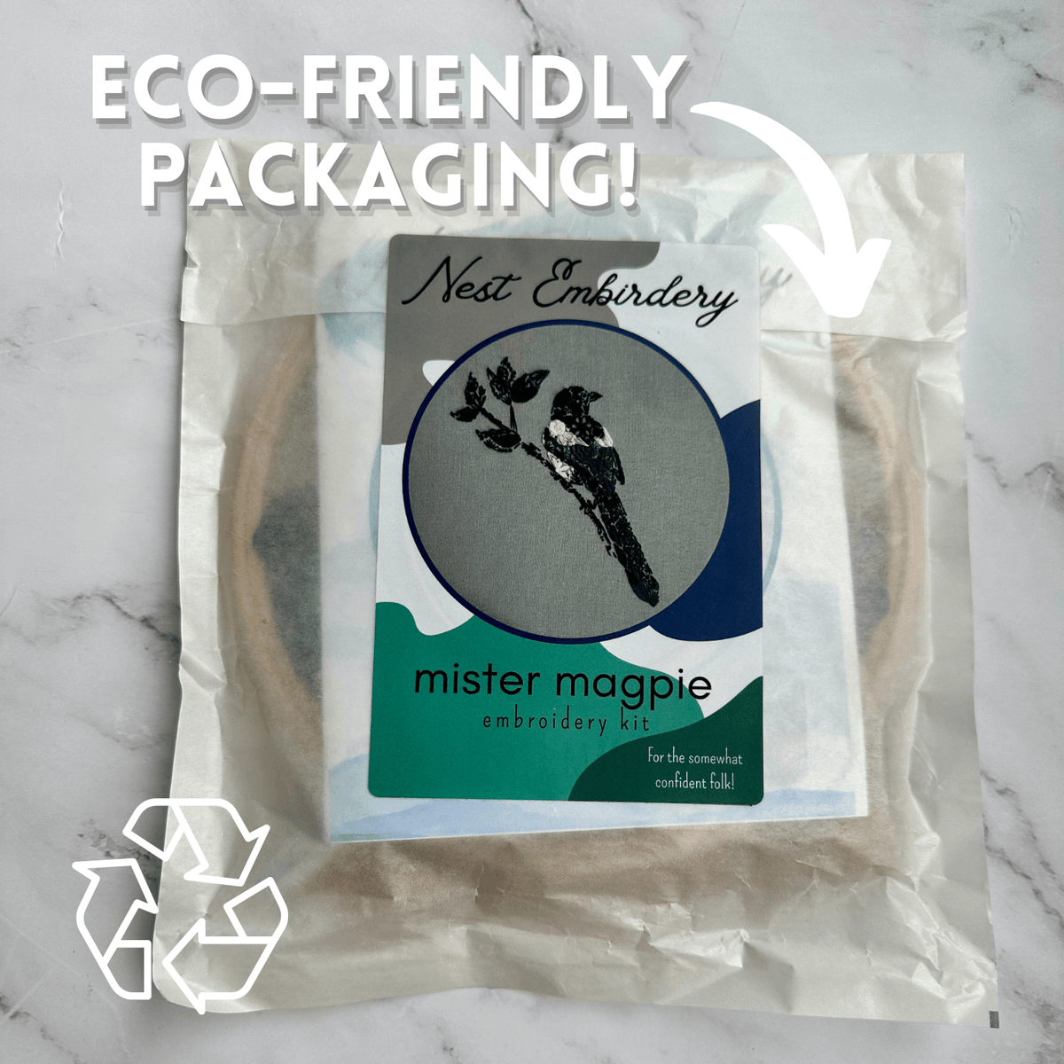 The 7 Beginner Mister Magpie Embroidery Kit by Nest Embirdery features a magpie bird design and eco-friendly packaging with a recycling symbol, displayed on a light marble surface.