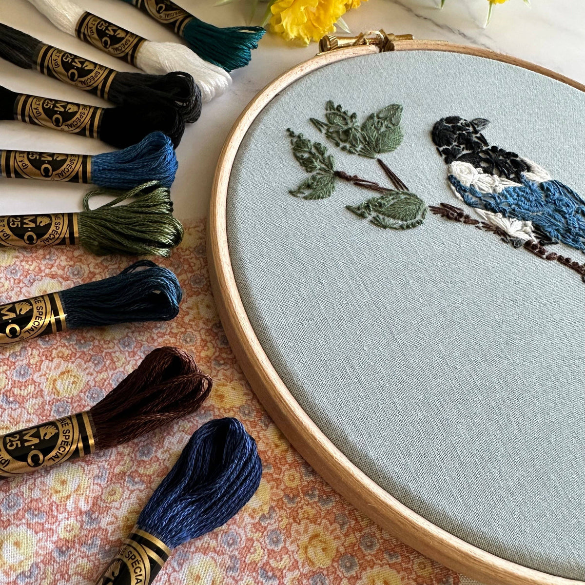 Nest Embirdery’s 7 Beginner Mister Magpie Embroidery Kit features a partially stitched magpie and branch on light fabric, framed by embroidery floss skeins atop a floral cloth, with yellow flowers adding charm—perfect for beginner stitchers.