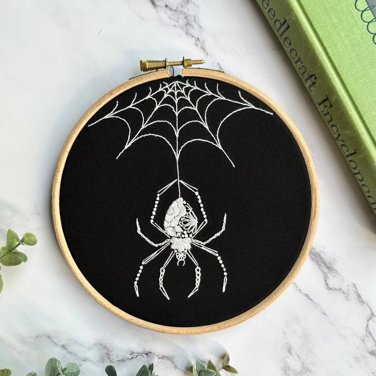 The Nest Embirdery 6 Beginner Orb Weaver Spider Embroidery Kit features an orb weaver spider stitched in white thread on black fabric, with green leafy details and a green book displayed on a marble surface.