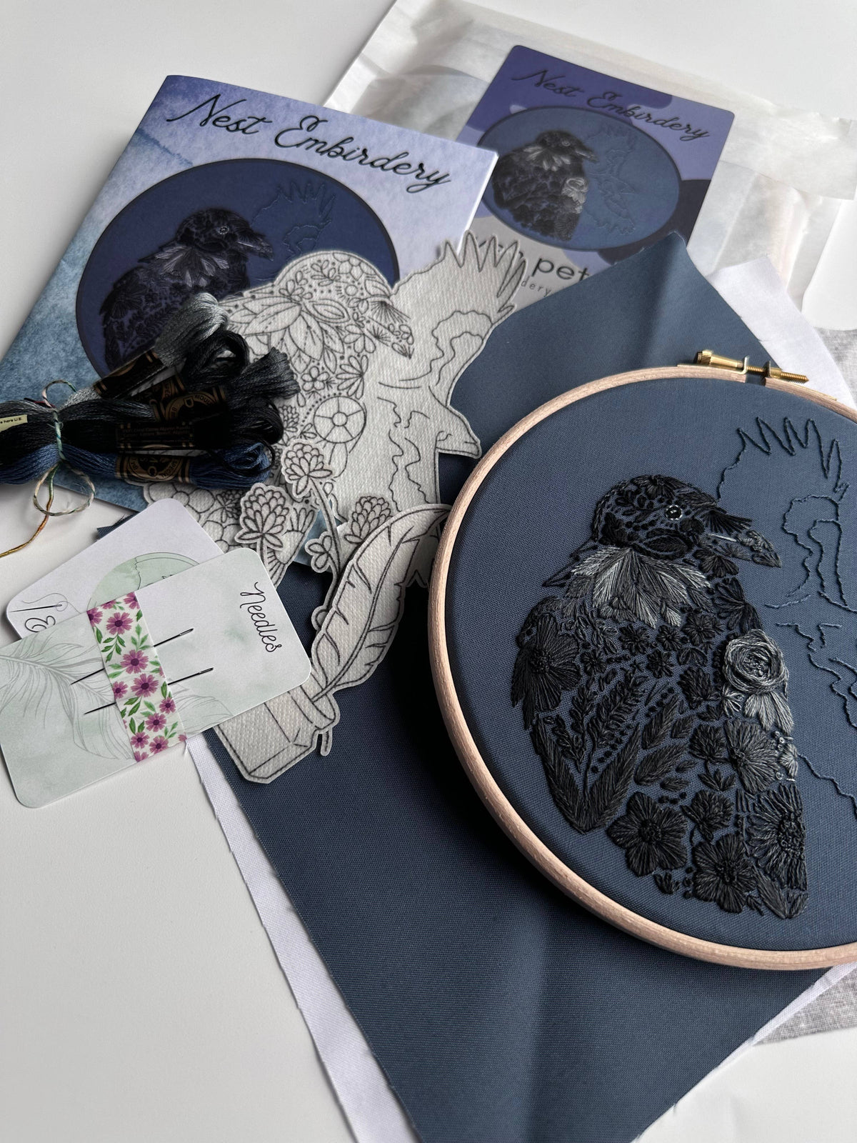 The 7 Beginner Raven Petals Embroidery Kit by Nest Embirdery features a black stitched raven on dark fabric in a wooden hoop, plus needle packs, embroidery floss, pattern outline, and branded packaging.