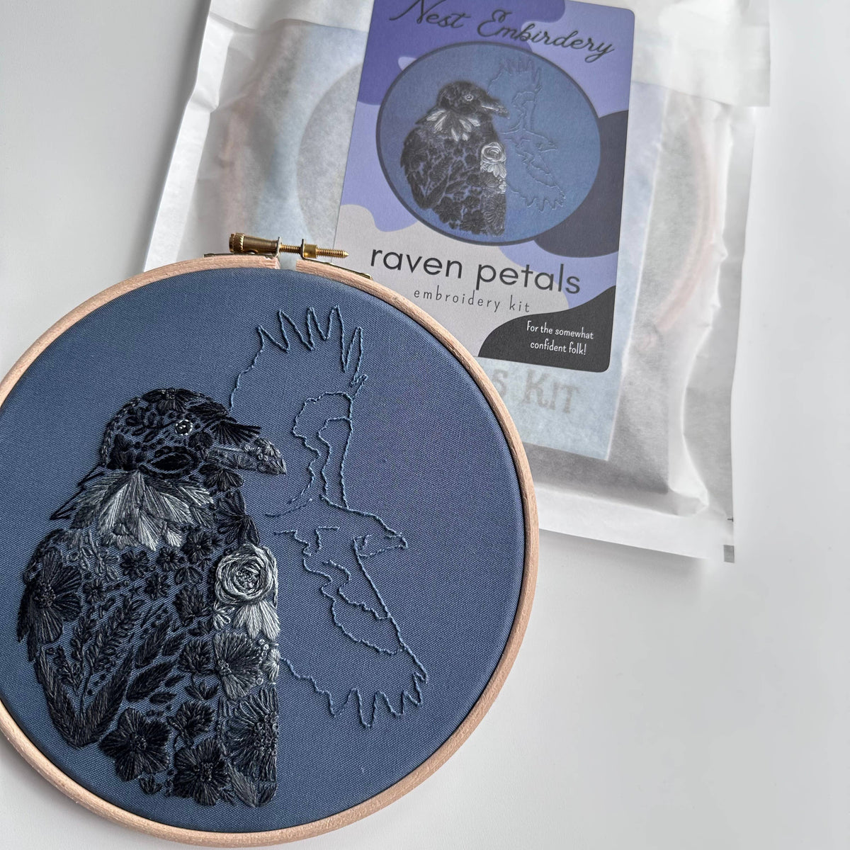 A finished 7 beginner raven embroidery from Nest Embirdery, featuring a black raven with floral accents and an outlined flying raven, is displayed next to its labeled Beginner Raven Petals Embroidery Kit packaging.