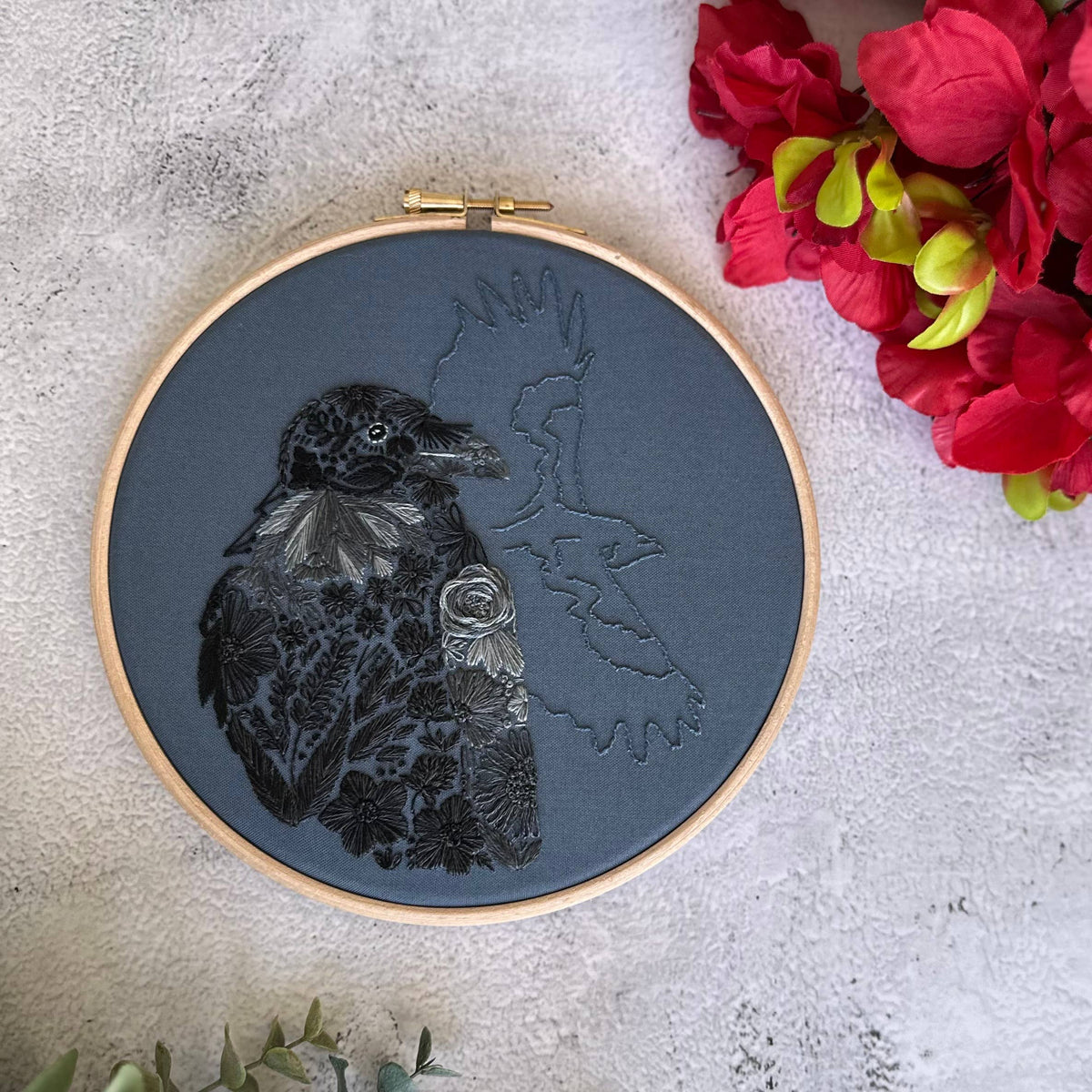 The Nest Embirdery 7 Beginner Raven Petals Embroidery Kit features a black raven with red and yellow floral patterns on textured gray fabric, plus a flying bird outline—perfect for beginners.