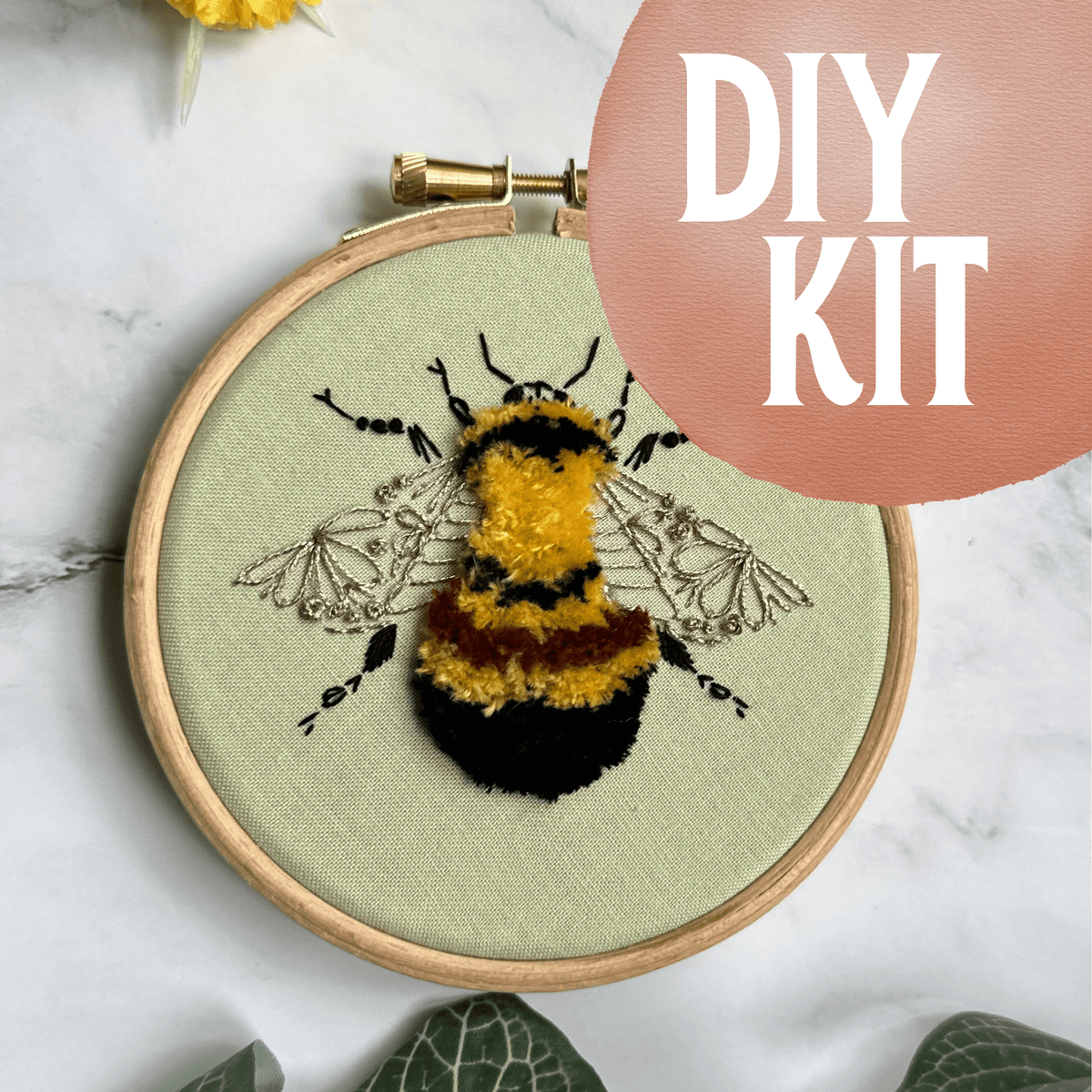 The Nest Embirdery 4 Beginner Rusty Patched Bumblebee Embroidery Kit features a textured bee on green fabric with black, yellow, and orange yarn. The DIY KIT label is in bold white on pink. Marble surface and green leaves are shown.