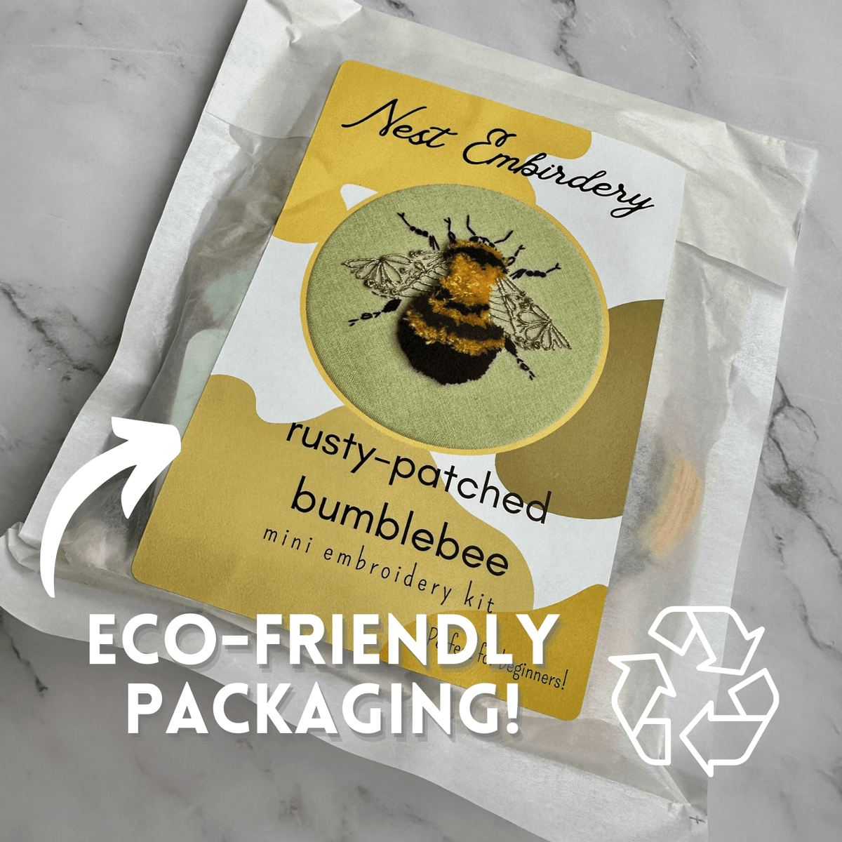 The 4 Beginner Rusty Patched Bumblebee Embroidery Kit by Nest Embirdery, featuring an endangered bee, comes in translucent paper. Text says ECO-FRIENDLY PACKAGING! with a recycling symbol and arrow directing attention to the package.
