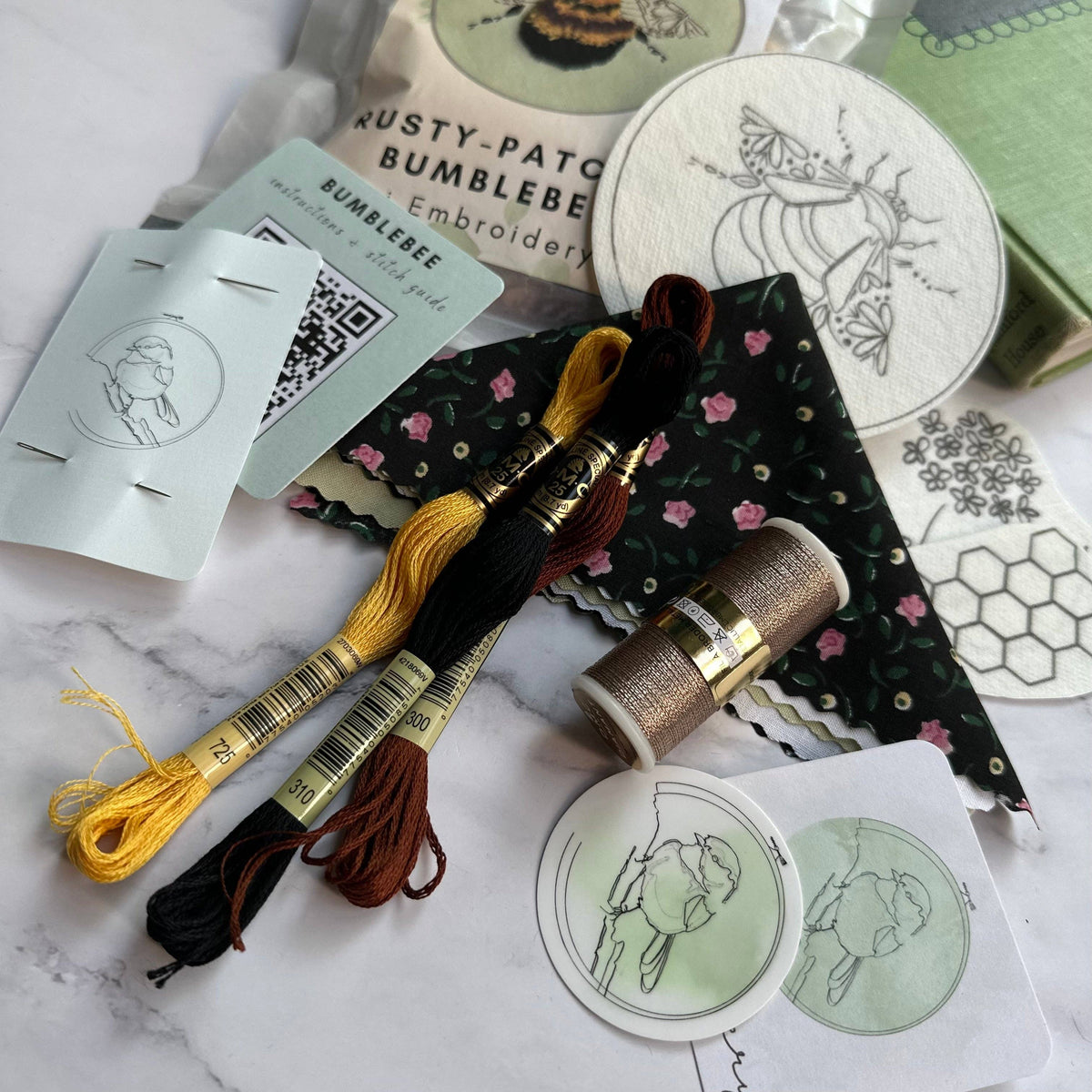Flat lay of the 4 Beginner Rusty Patched Bumblebee Embroidery Kit by Nest Embirdery, featuring bee-themed floss, metallic thread, fabric, patterns, stickers, and a book—all celebrating this endangered species.