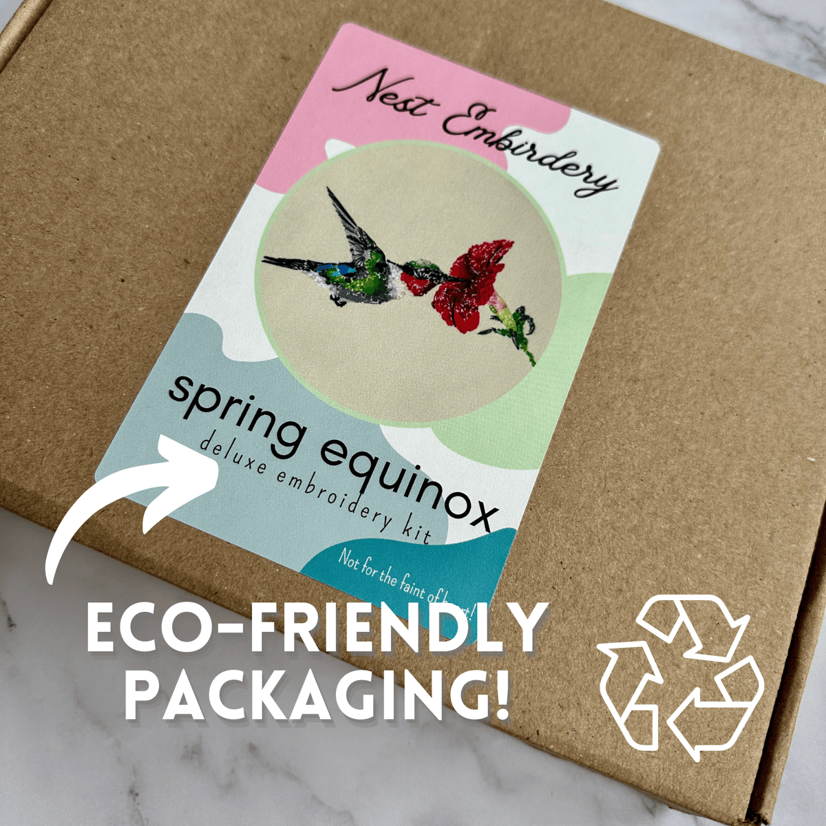 A brown box with a vibrant Nest Embirdery label displays two embroidered birds and floral hummingbirds. Text reads, 6 Spring Equinox Hummingbird Deluxe Embroidery Kit. Eco-friendly packaging! Recycling symbol shown.