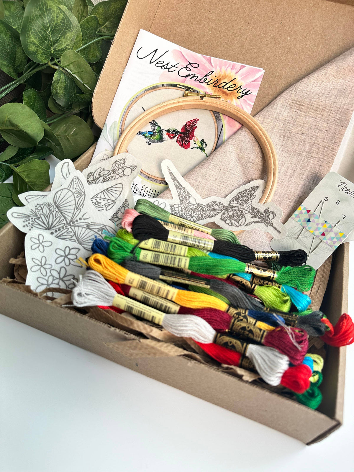 The Nest Embirdery 6 Spring Equinox Hummingbird Deluxe Embroidery Kit includes colorful threads, a wooden hoop, fabric, floral and butterfly Stick n Stitch designs, needles, instructions, and features green leaves beside the box.