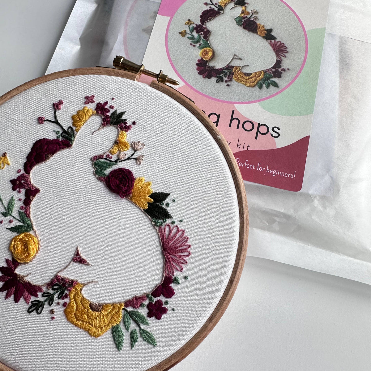 A 6 hoop displays a white fabric with a rabbit and floral embroidery from the Nest Embirdery Beginner Spring Hops Embroidery Kit. The partially visible kit package in the background features the same design and branding.