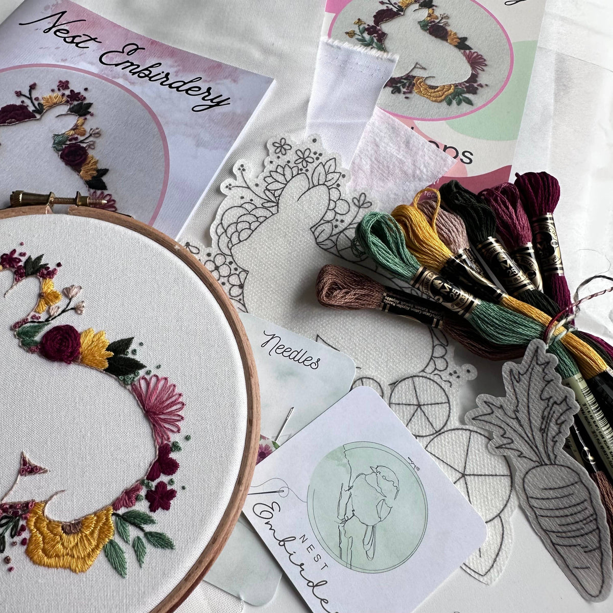 The Nest Embirdery 6 Beginner Spring Hops Embroidery Kit includes an embroidery hoop with a colorful floral design, pattern sheets, instruction cards, and embroidery floss bundles arranged on a white surface.