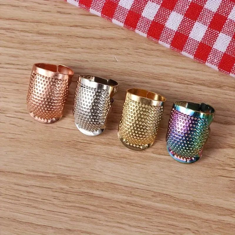 Four adjustable thimbles by Persephones Hearth in copper, silver, gold, and rainbow colors are lined up on a wooden surface next to red and white checkered fabric.