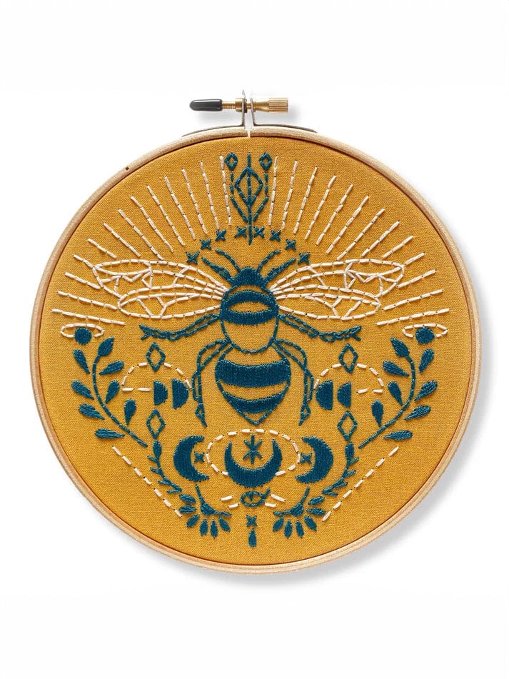 The *Late May Pre-Order* 6 Beginner Bee Embroidery Kit by Rikrack features a yellow fabric in a hoop with blue and white bee embroidery, floral patterns, crescent moons, stars, and radiating lines.