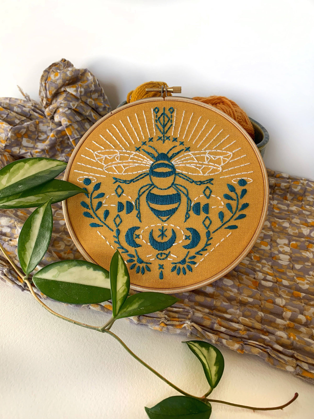 The *Late May Pre-Order* 6 Beginner Bee Embroidery Kit by Rikrack features a stylized blue and white bee with floral and geometric patterns on mustard fabric—ideal for beginners. The hoop is displayed on patterned fabric beside a leafy plant.