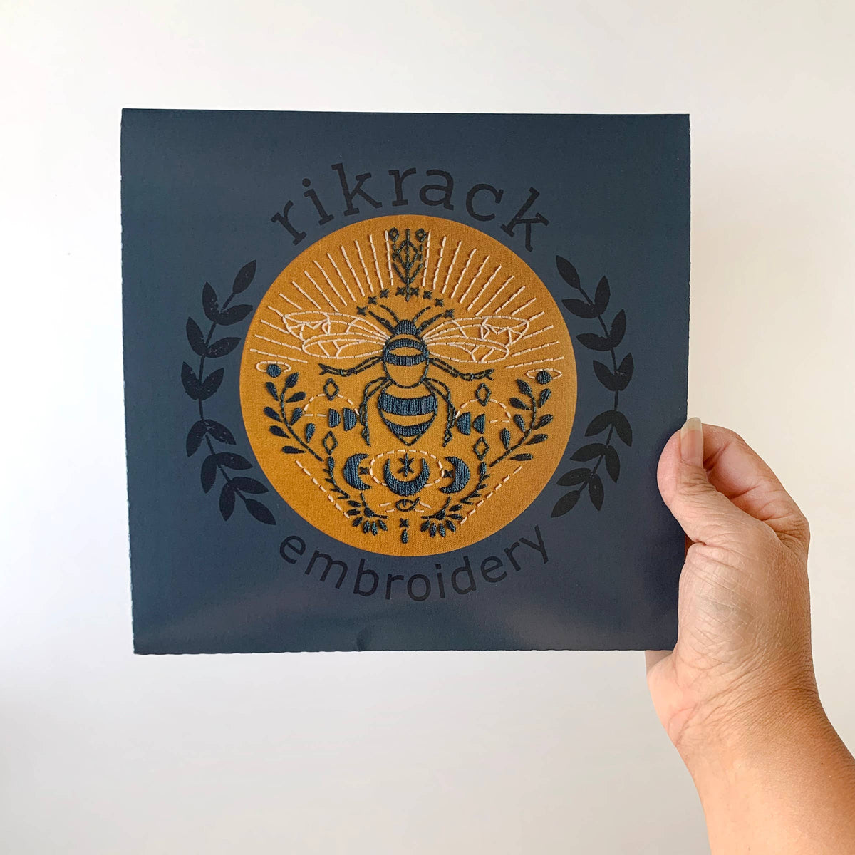 A hand holds a square dark blue card featuring the Rikrack *Late May Pre-Order* 6 Beginner Bee Embroidery Kit, showing an embroidered bee, foliage, and rays with “rikrack embroidery” encircling the motif—ideal for beginners.