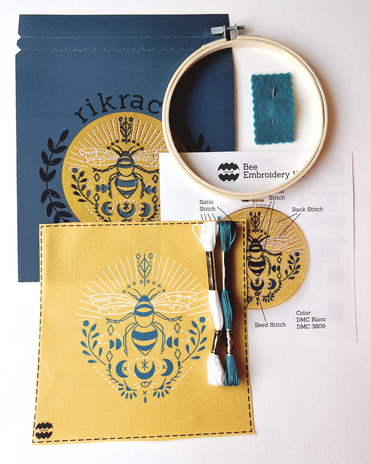 Flat lay of the *Late May Pre-Order* 6 Beginner Bee Embroidery Kit by Rikrack, with yellow bee design fabric, hoop, needle, 3 thread skeins, instructions, and blue Rikrack envelope—ideal for beginner crafters.