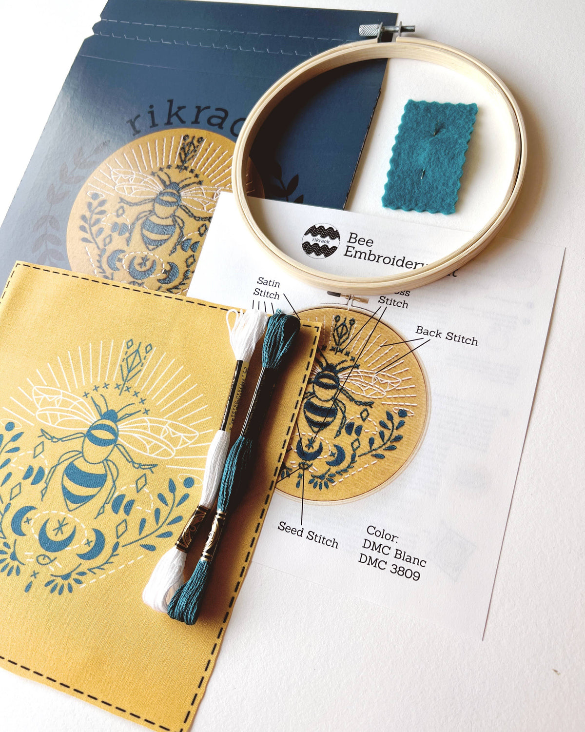 The Rikrack *Late May Pre-Order* 6 Beginner Bee Embroidery Kit includes printed fabric, thread, hoop, felt, and instructions featuring a blue bee on gold with floral designs—ideal for embroidery beginners.