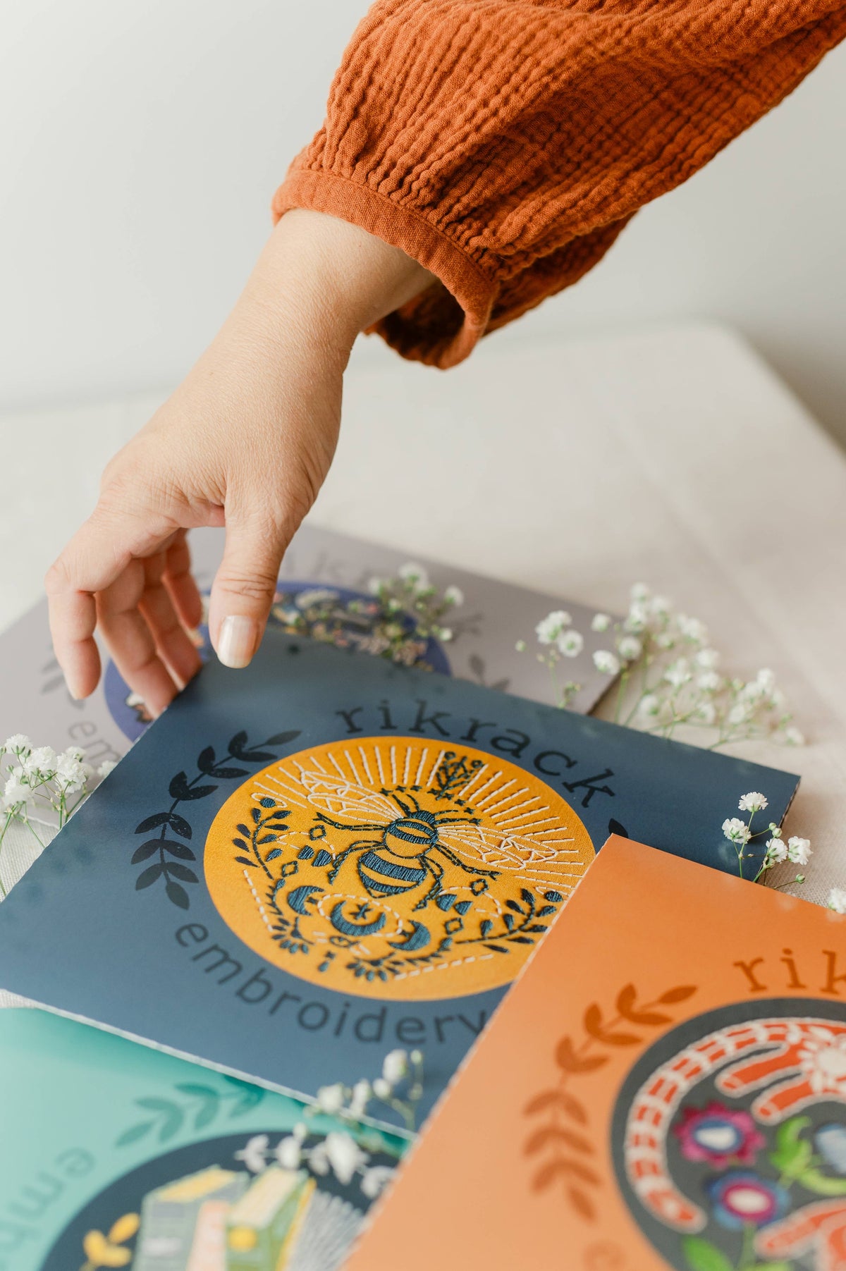 A hand in an orange sleeve reaches for *Late May Pre-Order* 6 Beginner Bee Embroidery Kits by Rikrack, displayed among small white flowers on a table. The kits feature colorful, intricate bee designs.