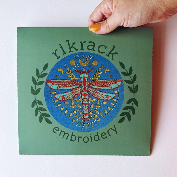 A hand holds a green square card displaying a dragonfly with lunar, star, and leafy branch details, along with Rikrack—the 6 Beginner Mystic Dragonfly Embroidery Kit, perfect for nature-loving beginners.