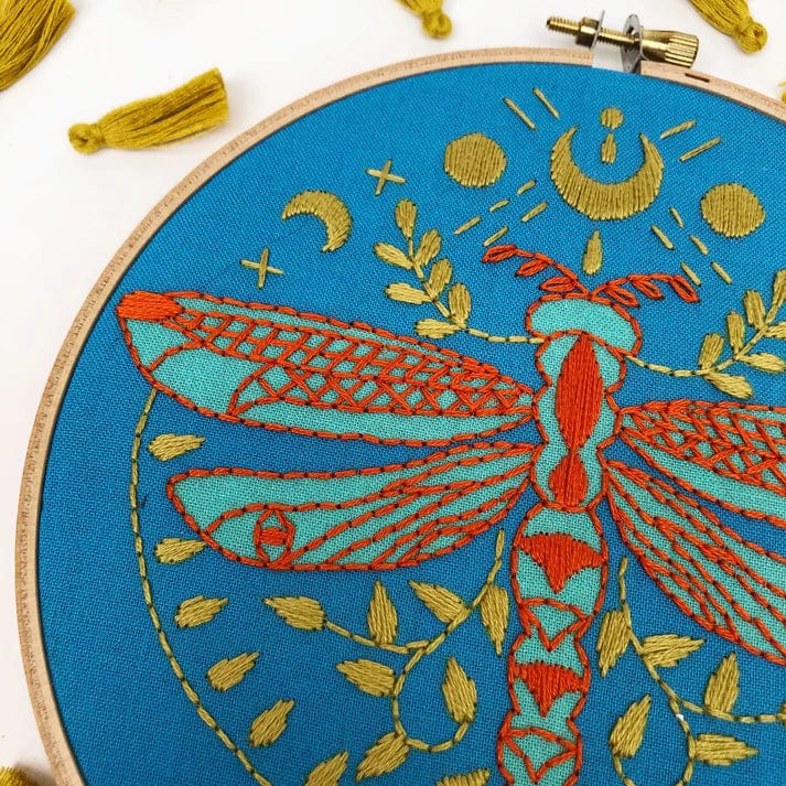 The Rikrack 6 Beginner Mystic Dragonfly Embroidery Kit features a red and gold dragonfly on blue fabric with gold leaves, moons, and stars. Framed in a wooden hoop with yellow tassels, its perfect for beginner stitchers.