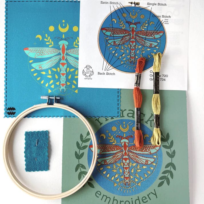 The Rikrack 6 Beginner Mystic Dragonfly Embroidery Kit features a colorful dragonfly design on a blue-green background with fabric, hoop, orange and yellow thread, needle, and instructions—perfect for beginner embroidery lovers.