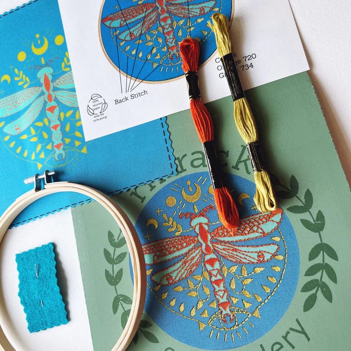 The Rikrack 6 Beginner Mystic Dragonfly Embroidery Kit includes a printed design, colorful threads, hoop, needle holder, and instructions—ideal for beginners to stitch a vibrant red dragonfly with blue and gold accents.