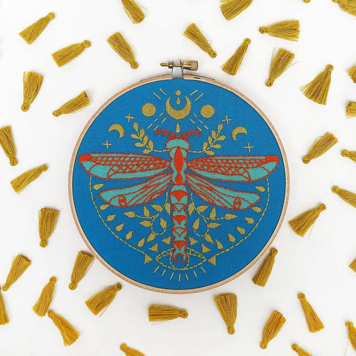 The Rikrack 6 Beginner Mystic Dragonfly Embroidery Kit features a colorful dragonfly with geometric and leaf designs on blue fabric, accented by gold tassels—perfect for beginners and displayed on a white background.