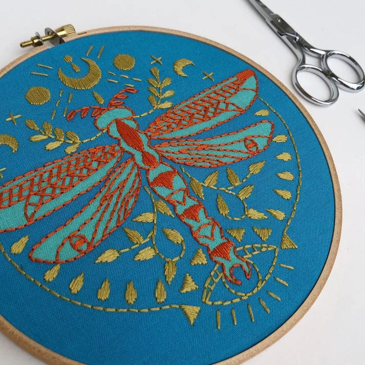 The Rikrack 6 Beginner Mystic Dragonfly Embroidery Kit features a blue hoop with red and gold dragonfly embroidery, gold moon and leaf motifs, plus included embroidery scissors and needle, all displayed on a white surface.