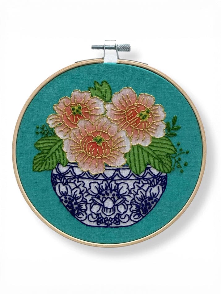The Rikrack 6 Beginner Peony Embroidery Kit features coral peonies and pink blooms with green leaves in a blue and white vase, stitched on teal fabric—a vibrant, charming addition to any craft collection.