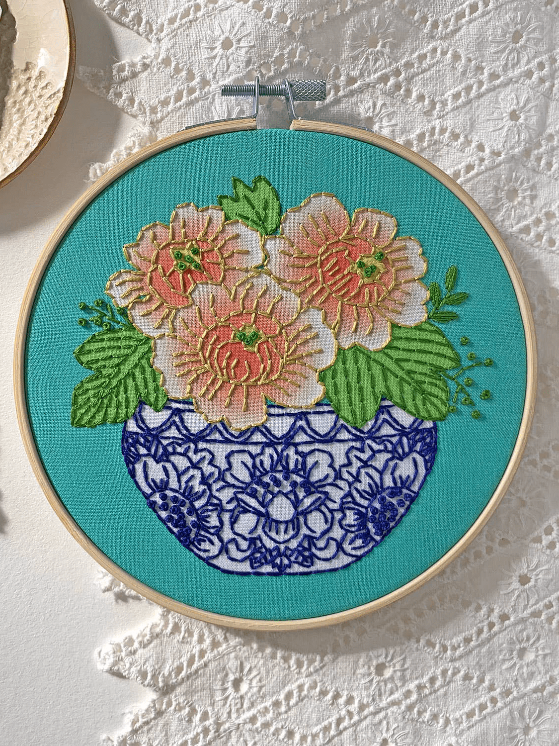 The Rikrack 6 Beginner Peony Embroidery Kit features pink and white flowers with green leaves in a blue and white vase on teal fabric, ideal for beginners to practice embroidery skills.