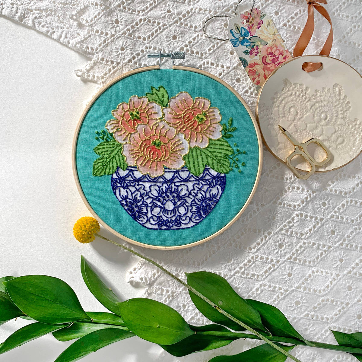 The Rikrack 6 Beginner Peony Embroidery Kit features a floral design of pink and coral peonies in a blue and white vase on teal fabric, with included scissors, thread, embroidery tips card, and decorative accents for beginners.