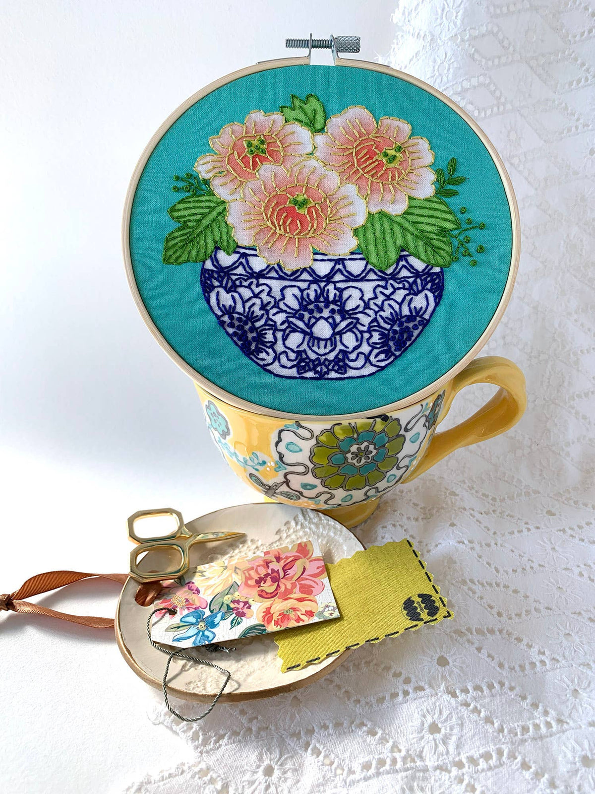The 6 Beginner Peony Embroidery Kit by Rikrack features a vibrant floral hoop with coral peonies atop a patterned teacup, surrounded by threads, scissors, and fabric pieces—perfect inspiration for beginners learning to embroider.