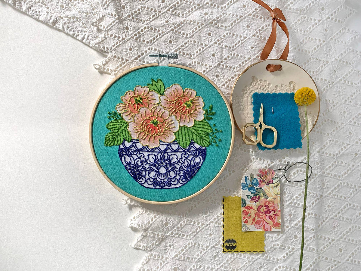 The Rikrack 6 Beginner Peony Embroidery Kit features Coral Peony Blooms in a blue vase on white lace fabric, with included scissors, needle, blue fabric, yellow flower, and floral paper—ideal for embroidery beginners.