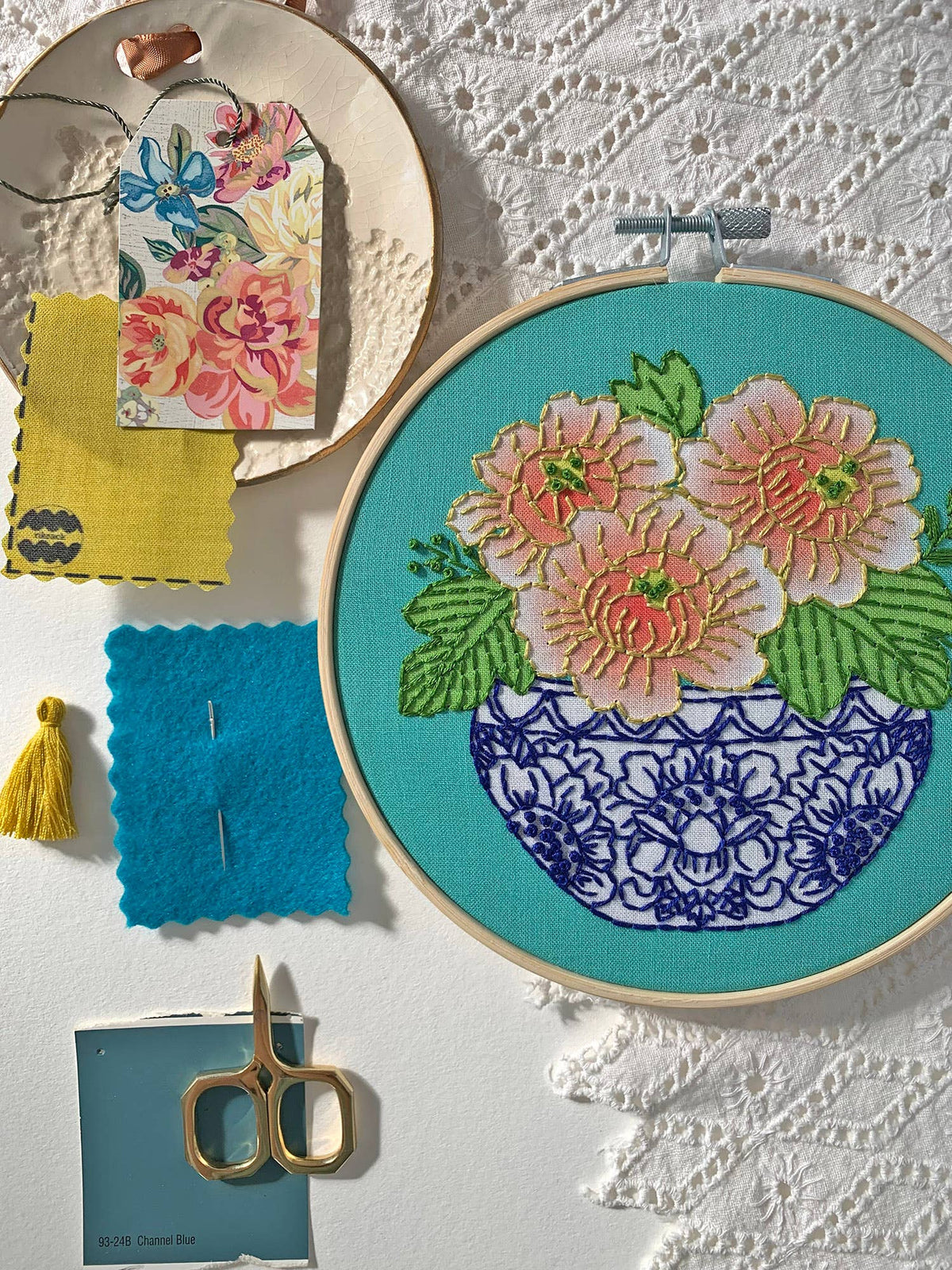The Rikrack 6 Beginner Peony Embroidery Kit features pink, peach, and coral peonies in a blue vase on teal fabric, surrounded by lace, swatches, a yellow tassel, gold scissors, a paint chip, and a floral tag. Perfect for beginners.