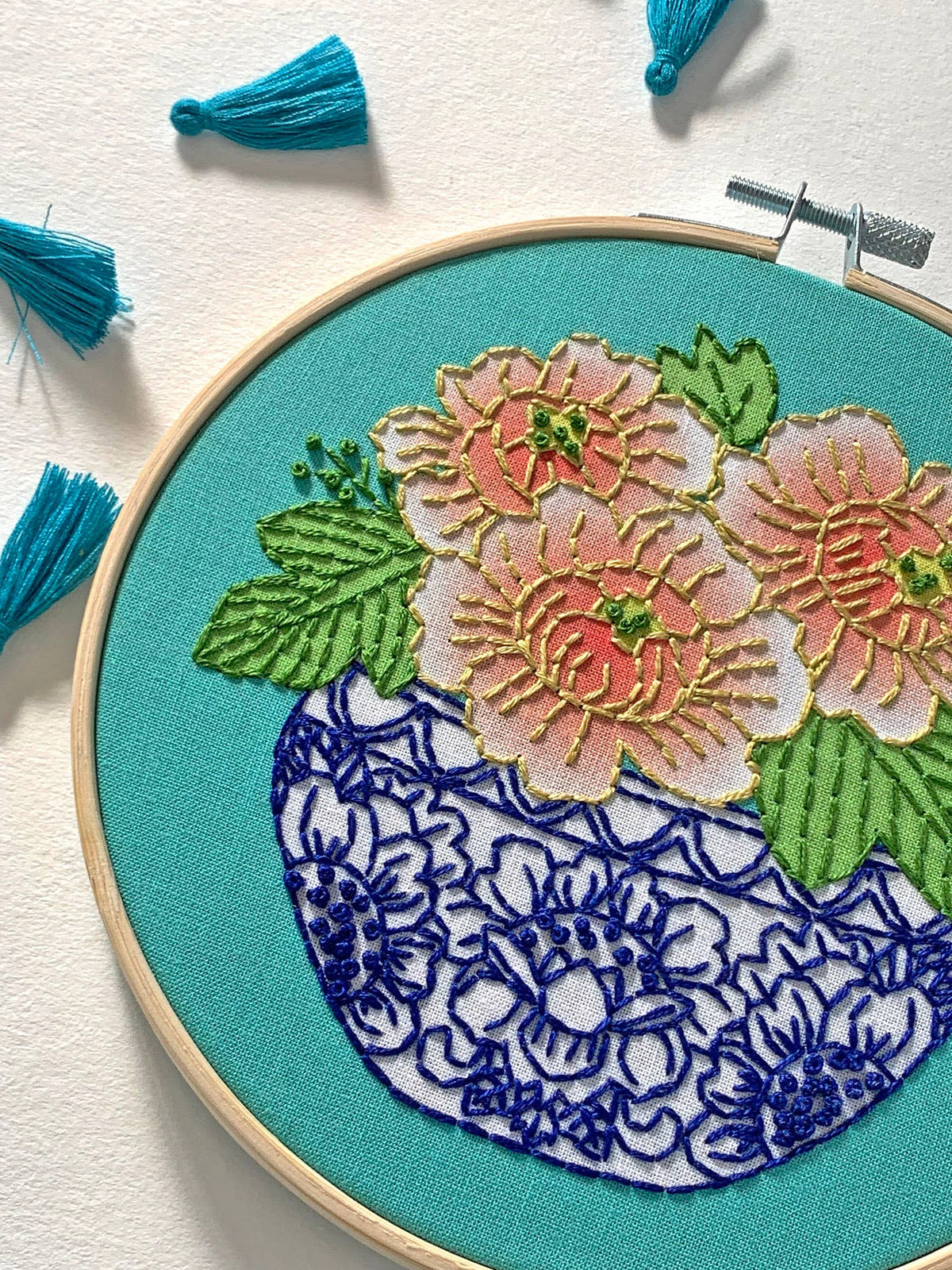 The Rikrack 6 Beginner Peony Embroidery Kit features Coral Peony Blooms with green leaves in a blue and white vase on turquoise fabric, accented by blue thread tassels—ideal for beginners.