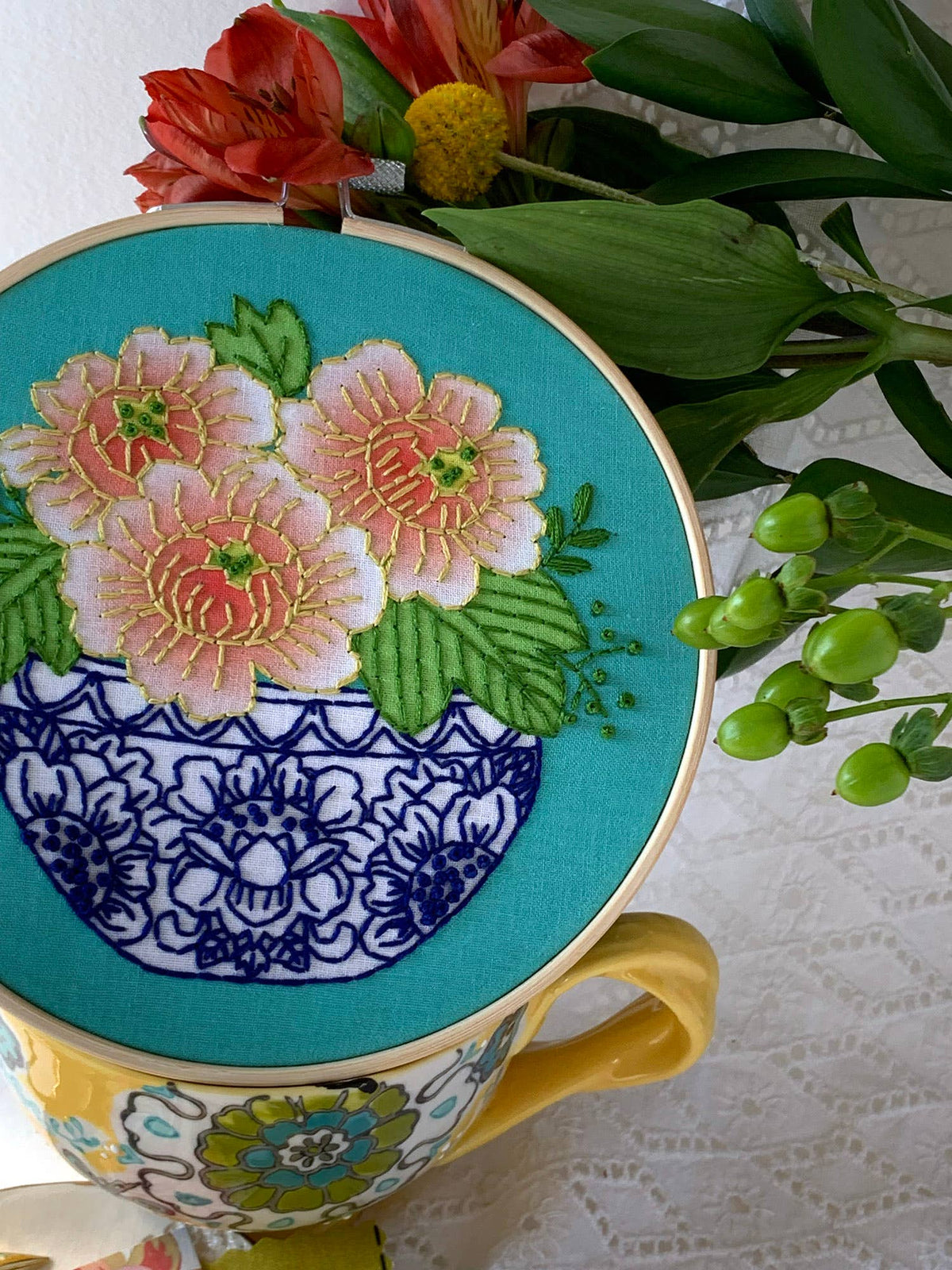 The Rikrack 6 Beginner Peony Embroidery Kit features pink and yellow flowers with green leaves in a blue vase on a teacup, plus nearby red and yellow peony blooms—perfect for beginners.