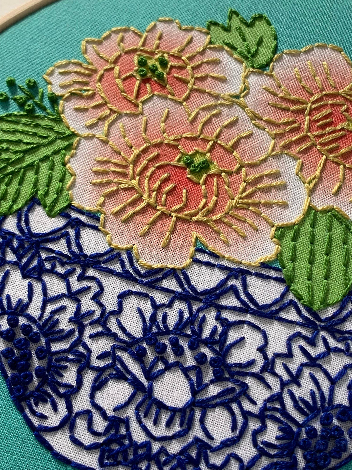 Close-up of hand embroidery with coral peony blooms, yellow flowers, and green leaves over dark blue lace-like stitching on light blue fabric—inspiration from Rikrack’s 6 Beginner Peony Embroidery Kit.
