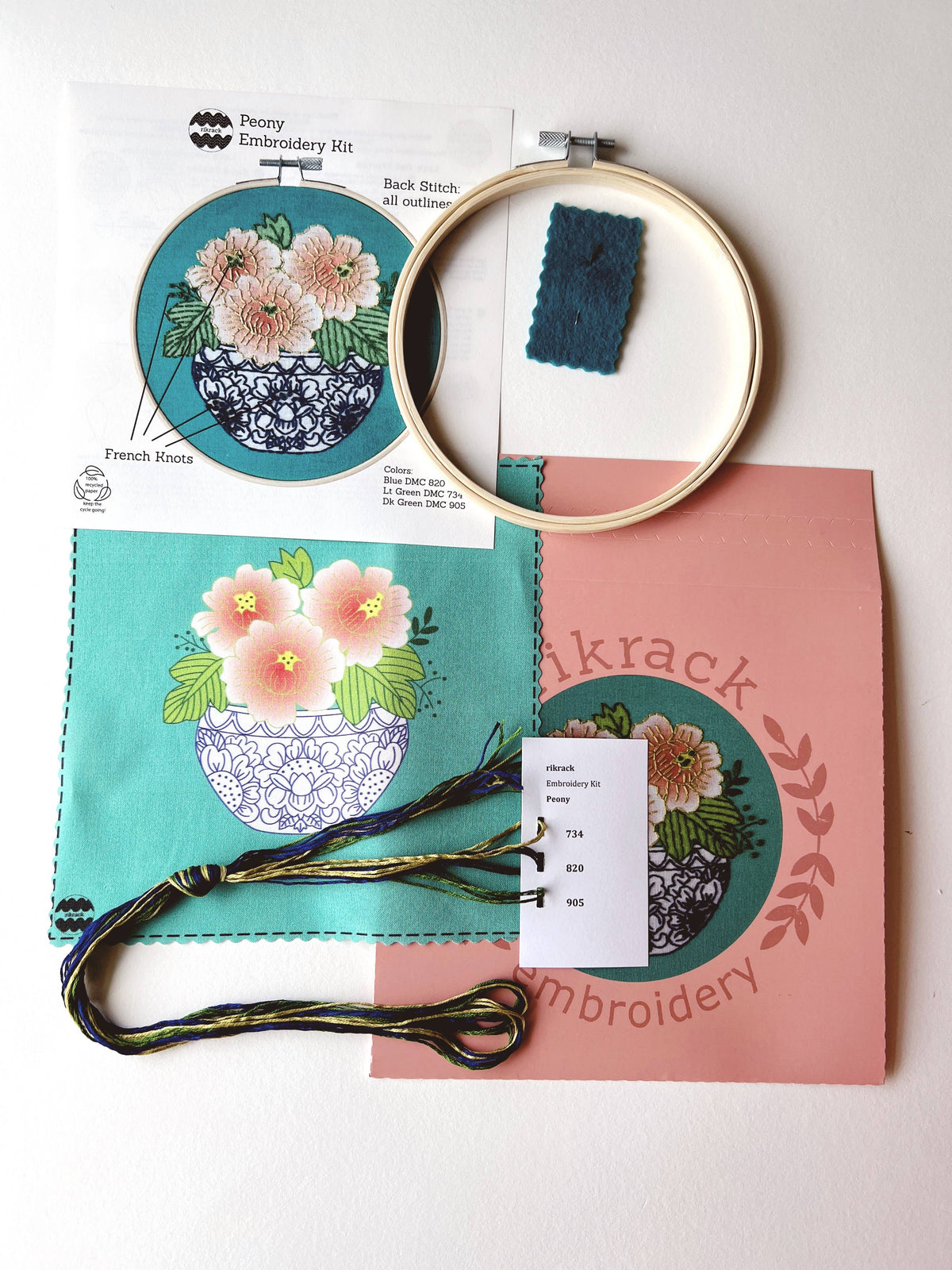 The Rikrack 6 Beginner Peony Embroidery Kit includes printed fabric with coral peony blooms, embroidery threads, wooden hoop, needle, instructions with tips, and a pink envelope labeled “Rikrack Embroidery.”.