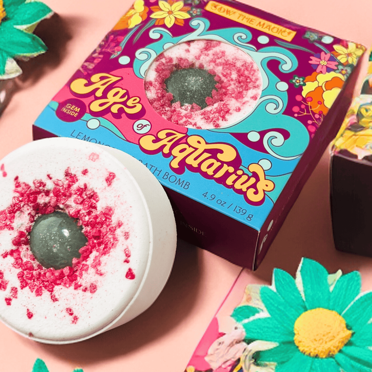 The Sow the Magic Age of Aquarius Lemongrass Bath Bomb with Aventurine, featuring pink and green accents, rests beside its vibrant floral box amid bright faux flowers—ideal for spiritual healing.