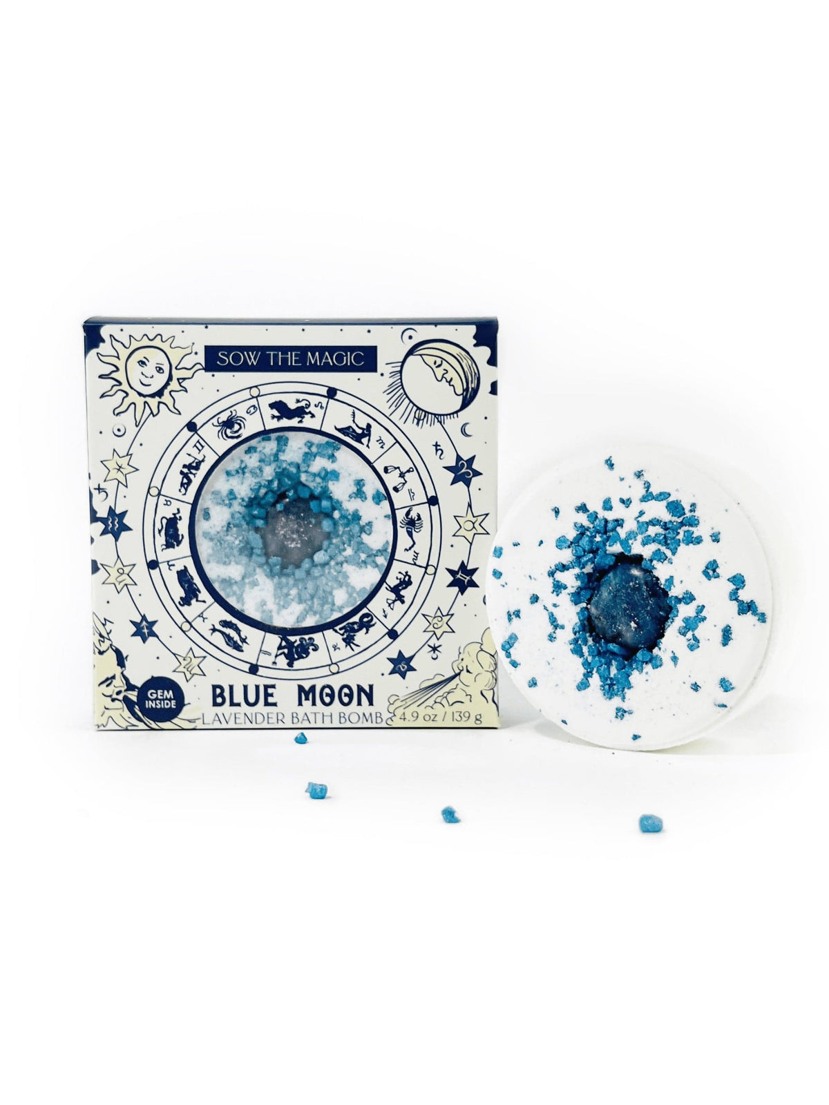 A blue and white Sow the Magic box labeled Blue Moon Lavender Bath Bomb with Sodalite stands beside a round, lavender-scented bath bomb topped with sodalite-like blue crystals, some scattered on a white surface.