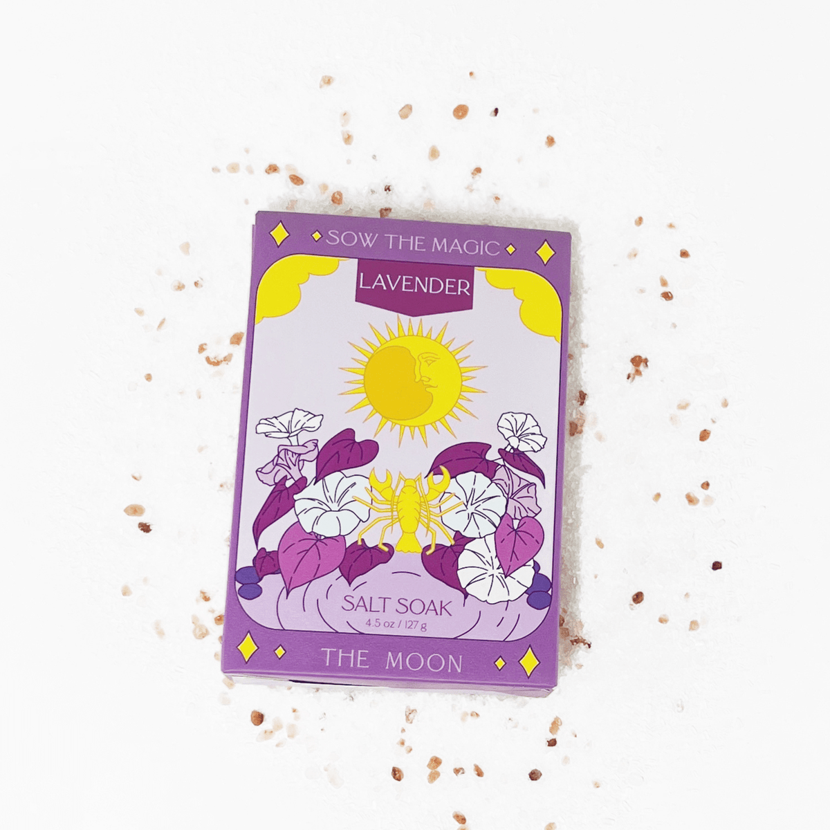 A packet of Sow the Magic The Moon Salt Soak in Lavender sits on a white surface with pink salt crystals. Its purple and yellow packaging features moonflowers, a sun illustration, and promotes mineral benefits.