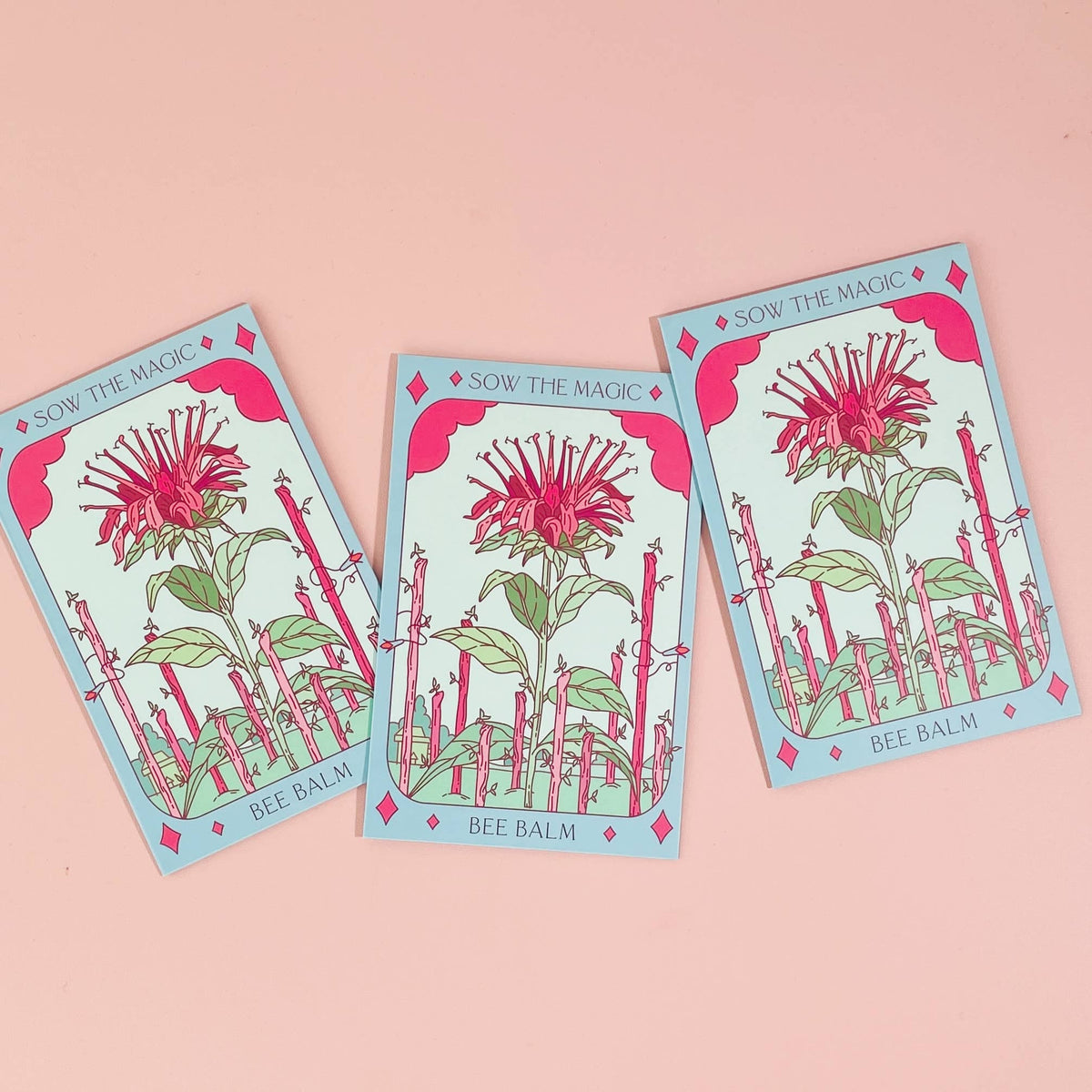Three Bee Balm Tarot Garden Seed Packets by Sow the Magic are displayed on a pink surface. Each packet features vibrant red and green Monarda didyma artwork, celebrating this pollinator-attracting flower.