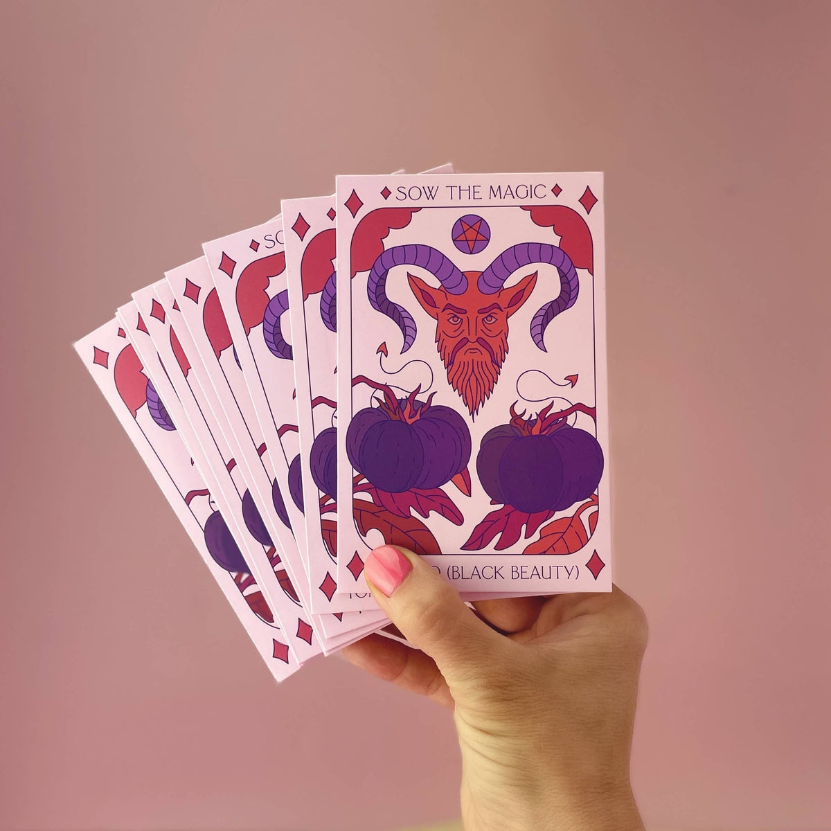 A hand with pink-painted nails holds a fan of illustrated cards. The top card, labeled Black Beauty Tomato Tarot Garden Seed Packet by Sow the Magic, features a goat head and purple pumpkins on a pink background.