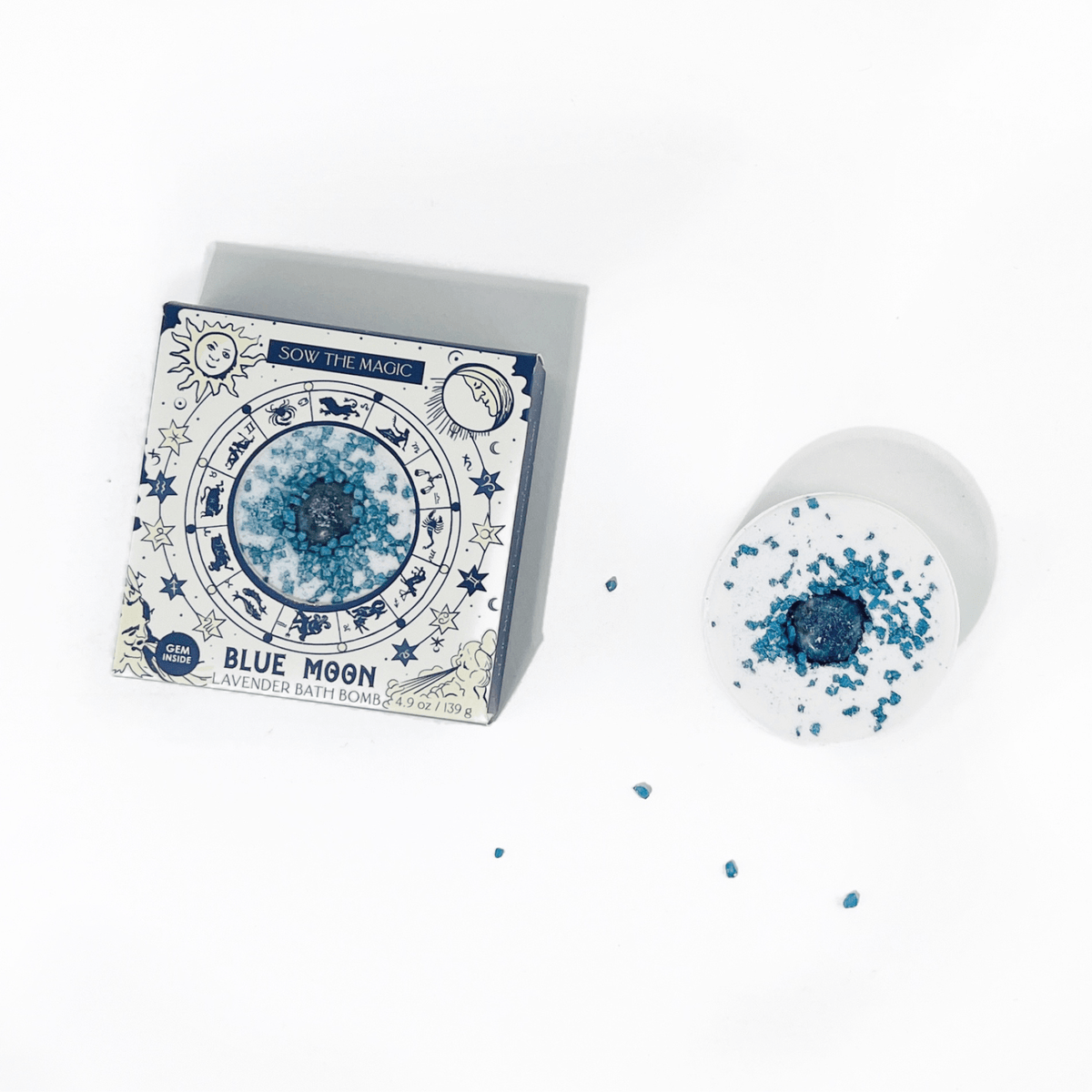 The Sow the Magic Blue Moon Lavender Bath Bomb with Sodalite sits beside its celestial-themed box, decorated with stars and moons, while sodalite-inspired blue crystals are scattered nearby.