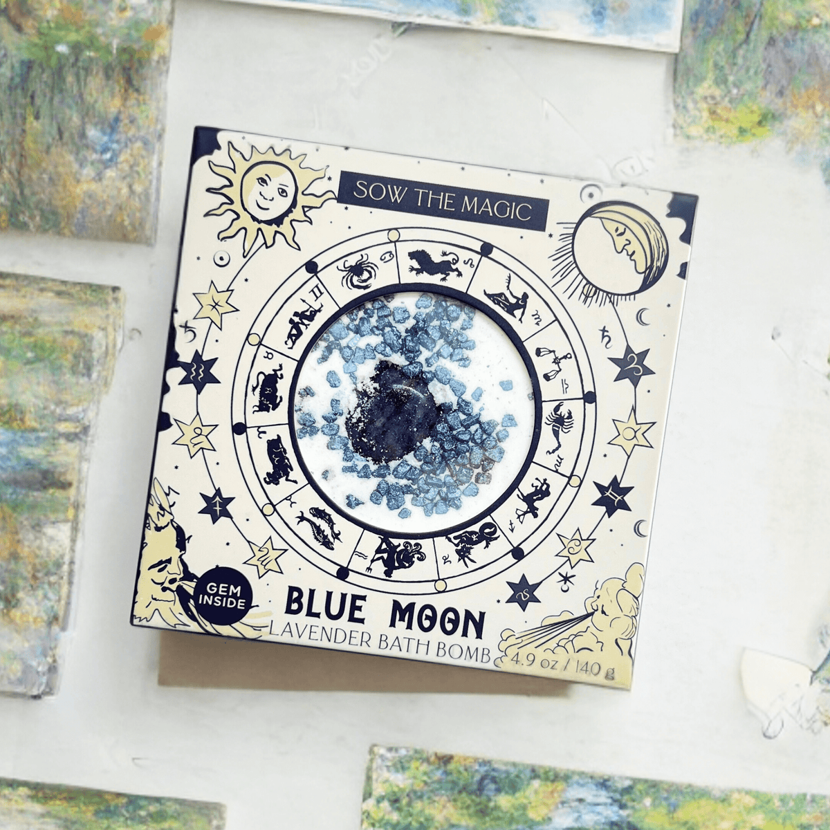The Sow the Magic Blue Moon Lavender Bath Bomb with Sodalite comes in a square box with zodiac and celestial designs, featuring a circular window revealing blue crystals and a hidden gemstone, plus packaging hints of “Gem Inside.”.