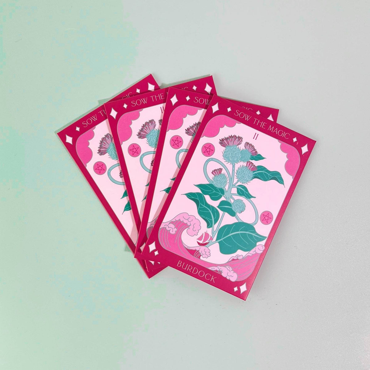 Four Burdock Tarot Garden Seed Packets by Sow the Magic, featuring illustrated detoxifying burdock with green leaves and purple flowers, are fanned out on a light gray surface.