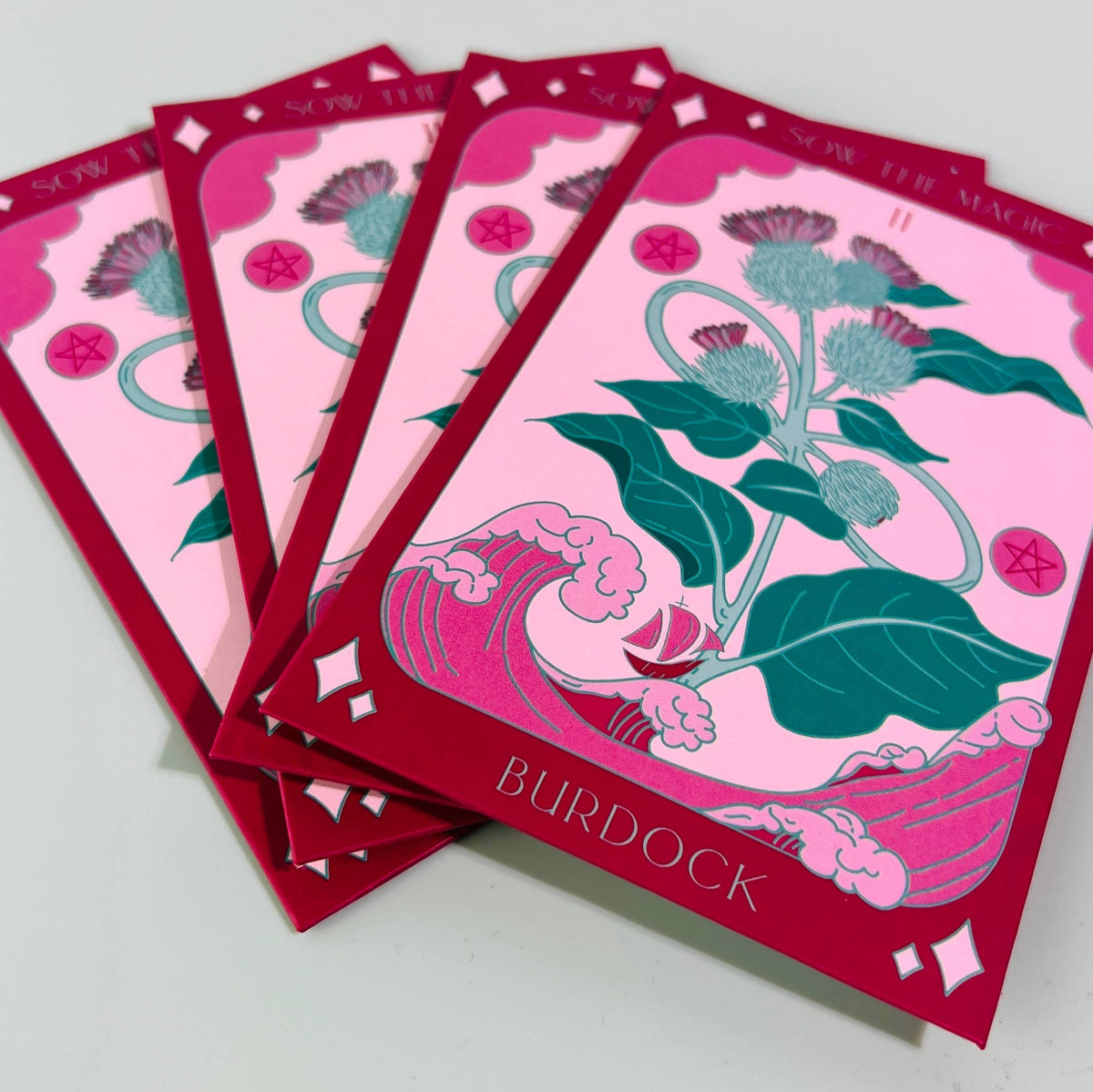 Four tarot-style cards display burdock plants with purple flowers and green leaves. Pink and red borders frame “BURDOCK” below, highlighting this detoxifying vegetable in the Sow the Magic Burdock Tarot Garden Seed Packet.